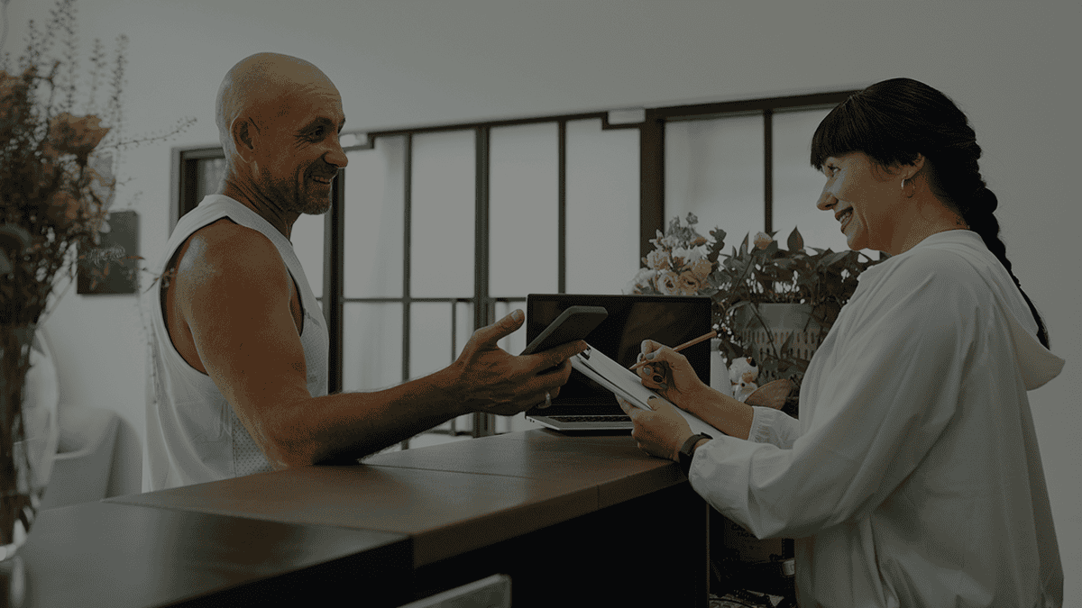 A man checks in at a Wodify gym reception desk, interacting with a smiling woman.
