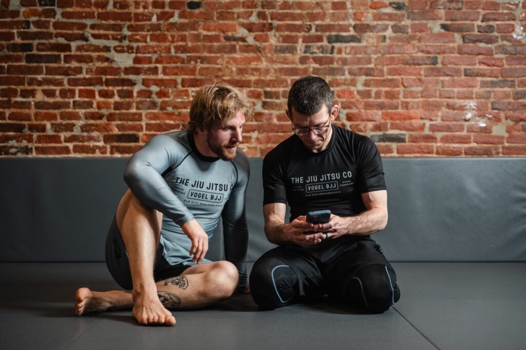 Two male athletes in Jiu Jitsu gis are looking at a phone on a gym mat, against a brick wall.