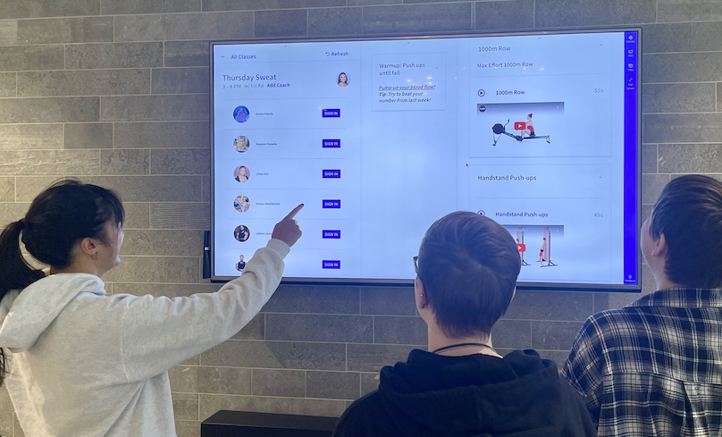 Three people view a Wodify gym class schedule and workout details on a large wall-mounted screen.